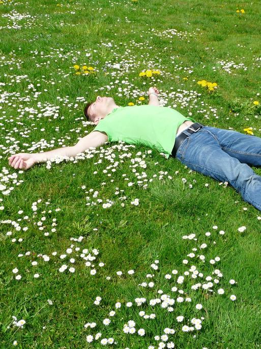 Ein Mann in Jeans und grünem Shirt liegt ausgestreckt mit dem Rücken auf einer blühenden Wiese und schläft.