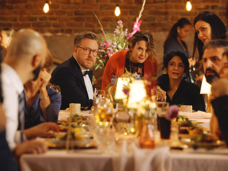 Filmszene aus Feste Freunde. Zu sehen sind Tom Trambow, Natalie (Jasmin Shakeri) und Maya (Katia Fellin) mit den Frischvermählten Rolf (Nicholas Ofczarek) und Dina (Pegah Ferydoni) an einer festlichen Tafel.