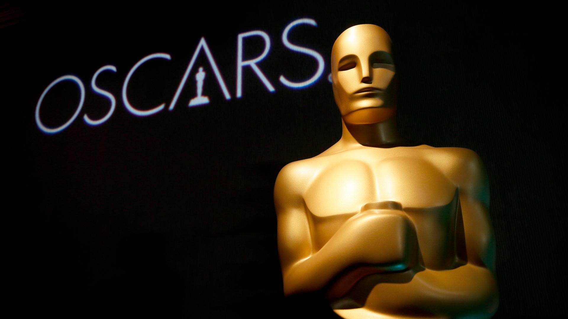 Das Bild zeigt eine große Oscar-Figur. Im Hintergrund steht auf einer schwarzen Wand "Oscars".