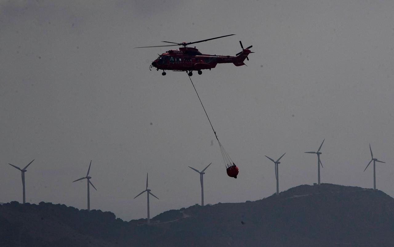 Ein Hubschrauber fliegt vor dem Hintergrund von Windrädern mit einem Behälter Wasser unter sich hängend über eine Landschaft.