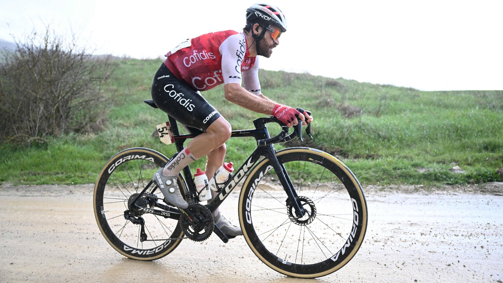 Zu sehen ist Radprofi Simon Geschke, während er auf einer nassen Straße bergan fährt.