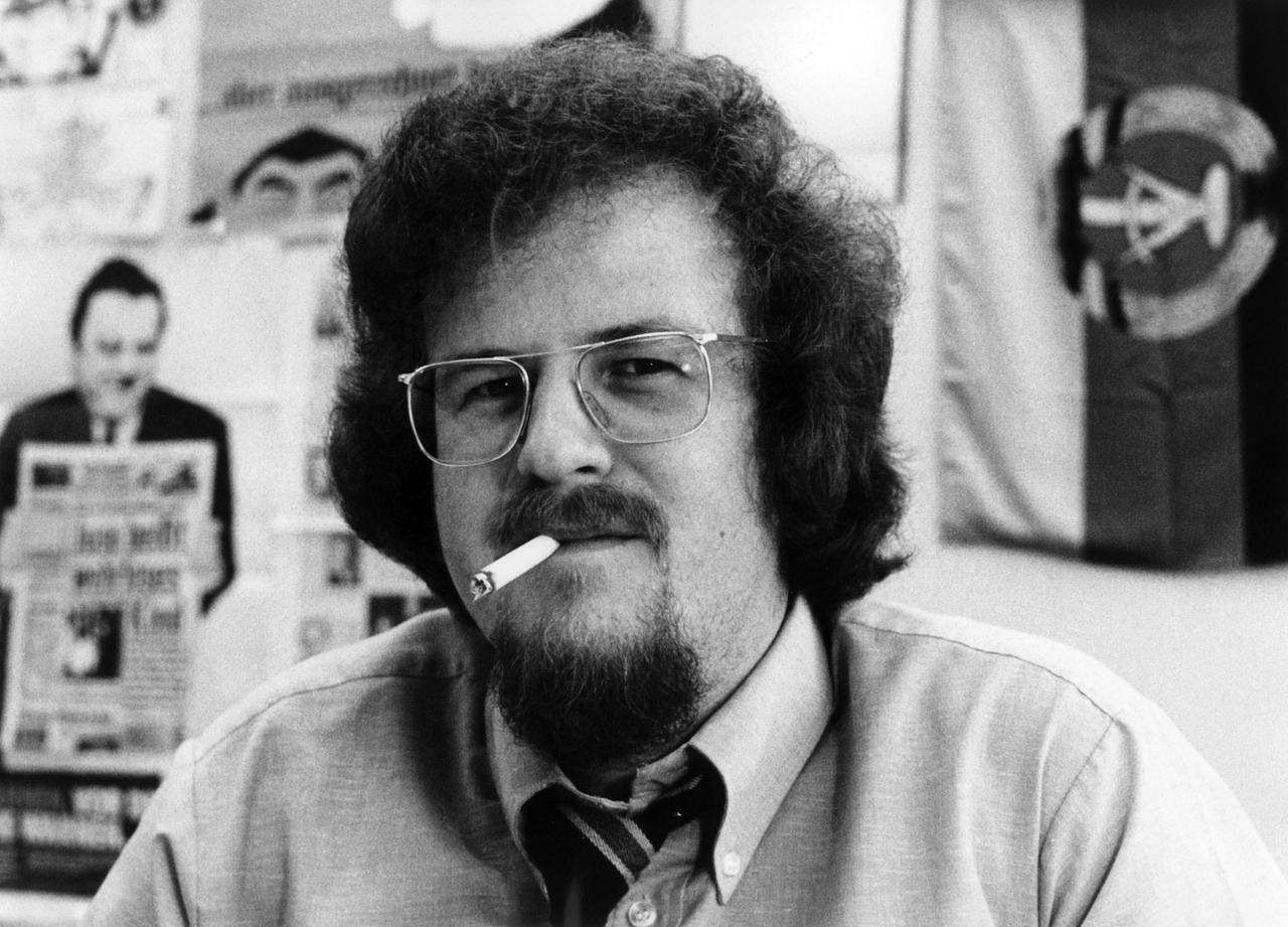 Jörg Mettke. Ein Mann mit lockigen Haaren und Brille sitzt in einem Büro und hat eine brennende Zigarette im Mund. Im Hintergrund hängen an der Wand eine DDR-Flagge und ein Bild von Franz Josef Strauß. 