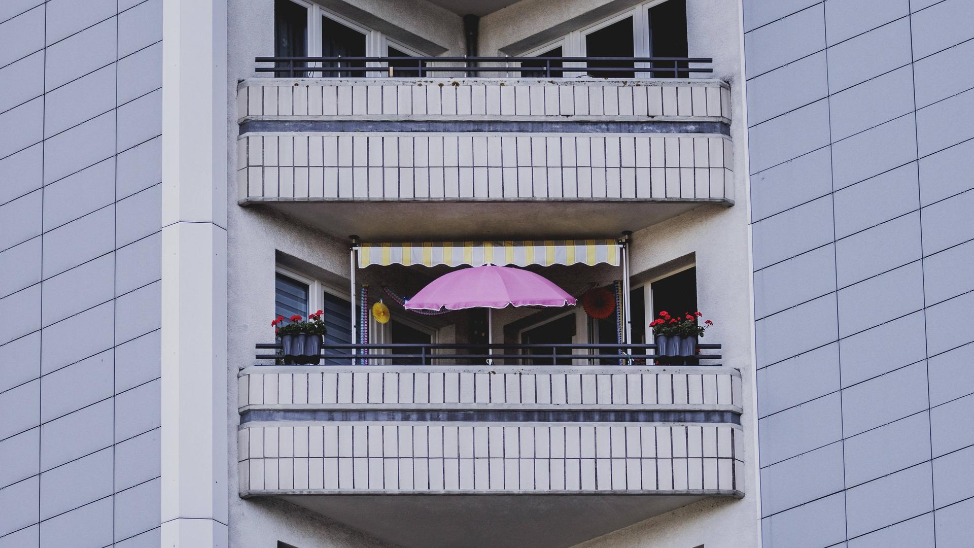 Das Foto zeigt einen pinkfarbenen Sonnenschirm auf dem Balkon eines Plattenbaus mit grauer Fassade in Berlin. Das Foto zeigt einen pinkfarbenen Sonnenschirm auf dem Balkon eines Plattenbaus mit grauer Fassade in Berlin.