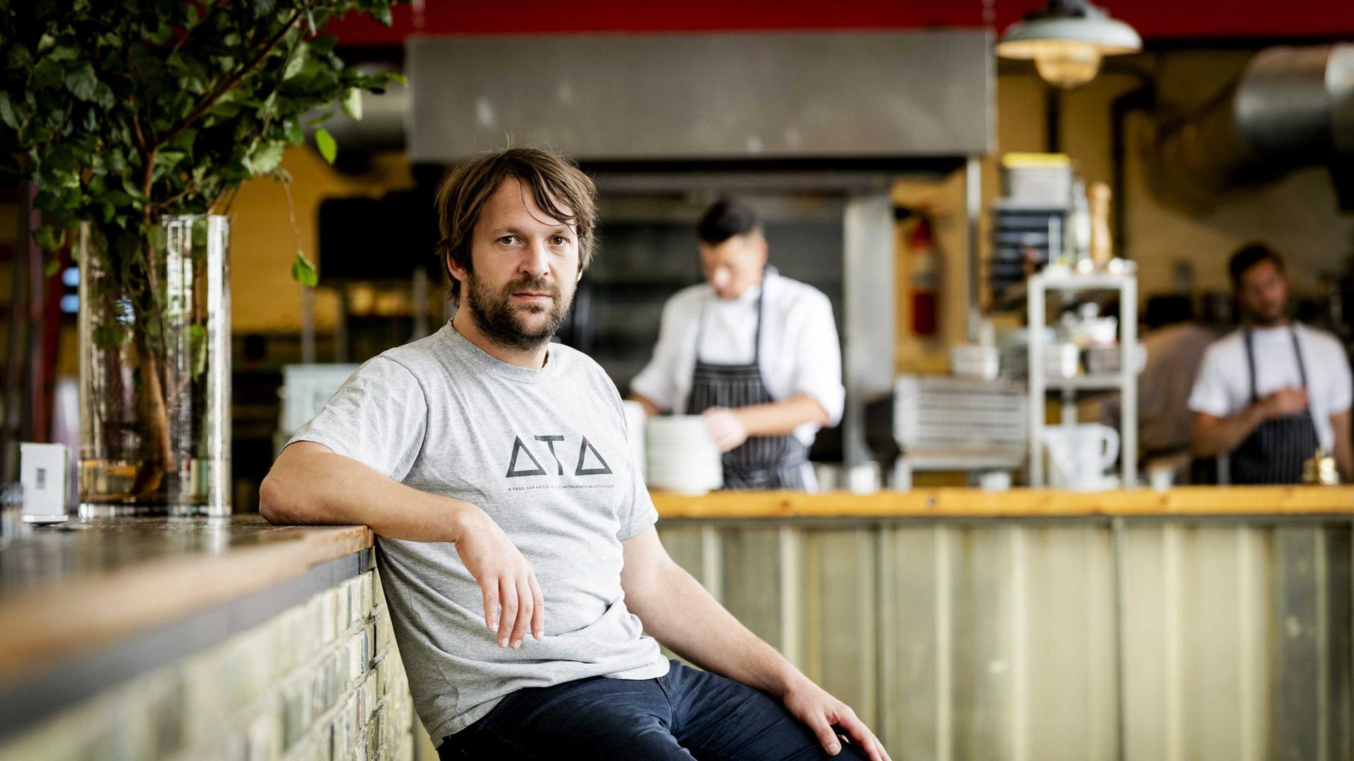 René Redzepi. Ein Mann mittleren Alters sitzt in dunklen Jeans und grauem T-Shirt am Tresen eines Restaurants.