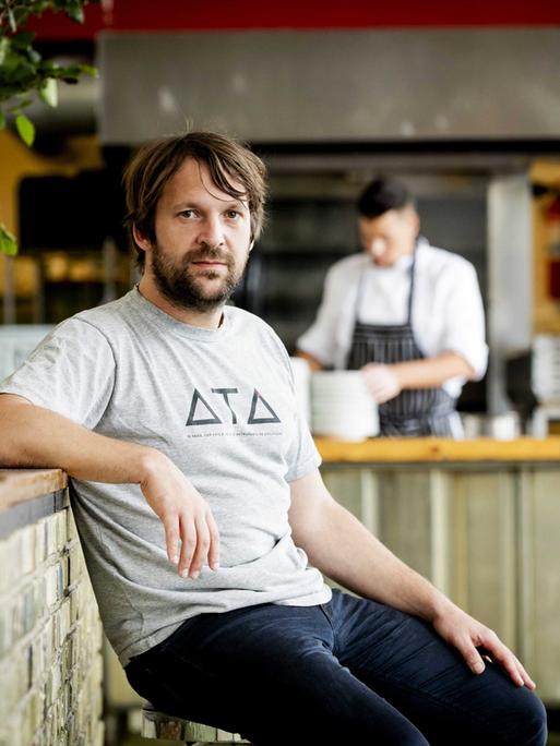 René Redzepi. Ein Mann mittleren Alters sitzt in dunklen Jeans und grauem T-Shirt am Tresen eines Restaurants.