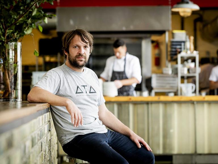 René Redzepi. Ein Mann mittleren Alters sitzt in dunklen Jeans und grauem T-Shirt am Tresen eines Restaurants.