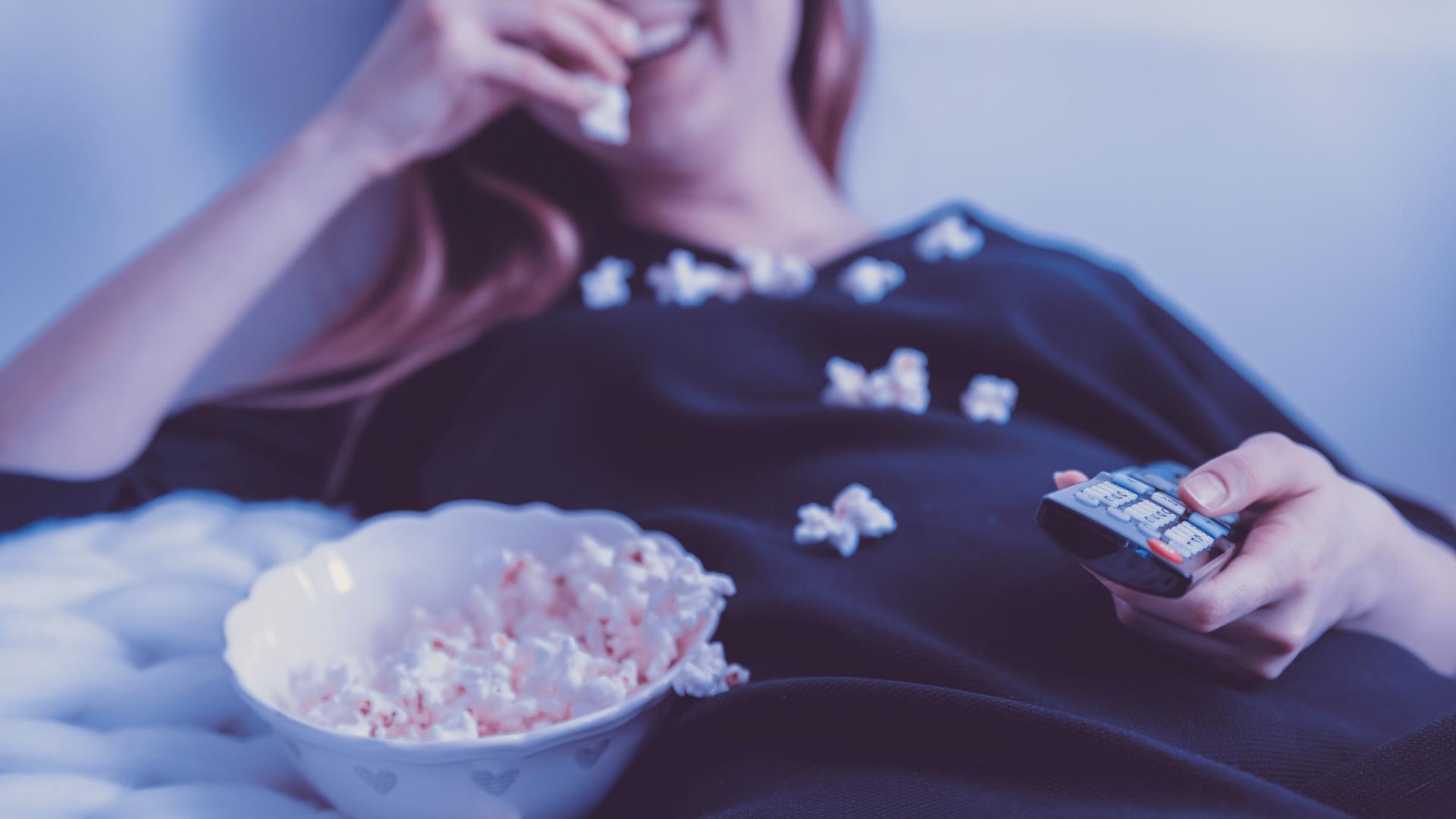 Eine Frau liegt mit einer Schale Popcorn und einer Fernbedienung auf dem Sofa.