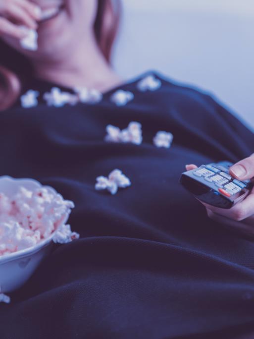 Eine Frau liegt mit einer Schale Popcorn und einer Fernbedienung auf dem Sofa.