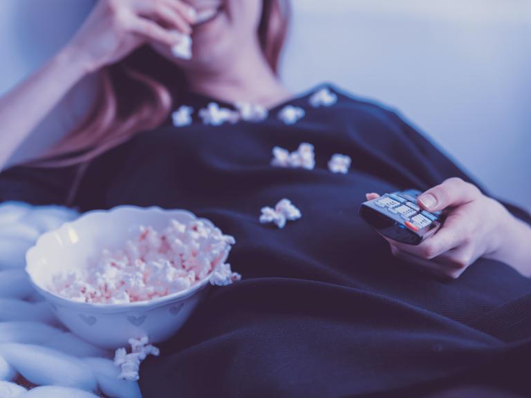 Eine Frau liegt mit einer Schale Popcorn und einer Fernbedienung auf dem Sofa.