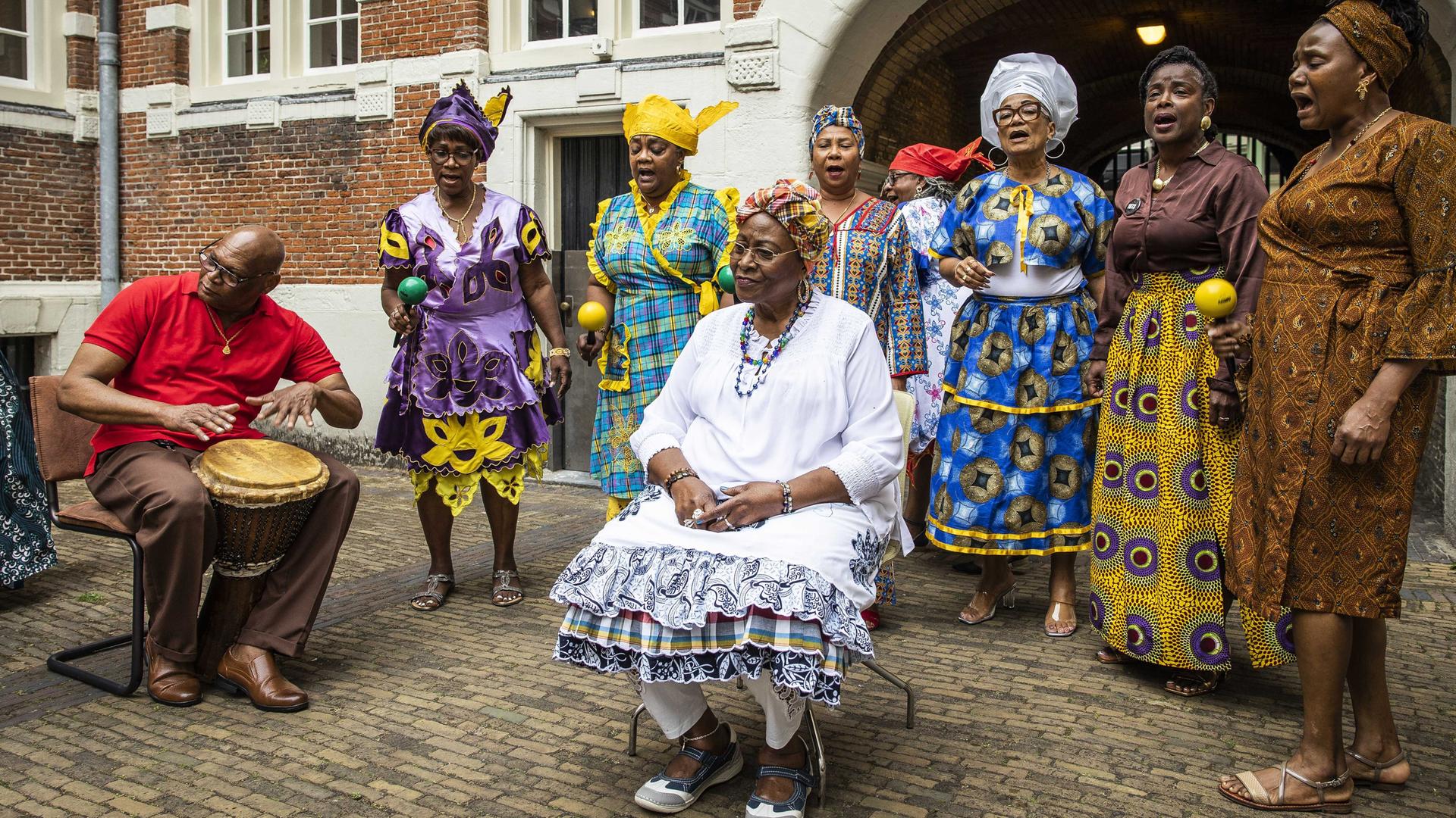 Menschen in farbenfrohen Kleidern singen in einem Hof vor einem Backsteingebäude.