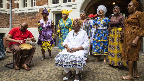 Menschen in farbenfrohen Kleidern singen in einem Hof vor einem Backsteingebäude.