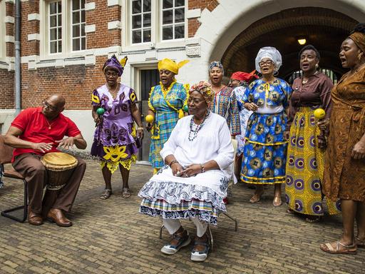 Menschen in farbenfrohen Kleidern singen in einem Hof vor einem Backsteingebäude.
