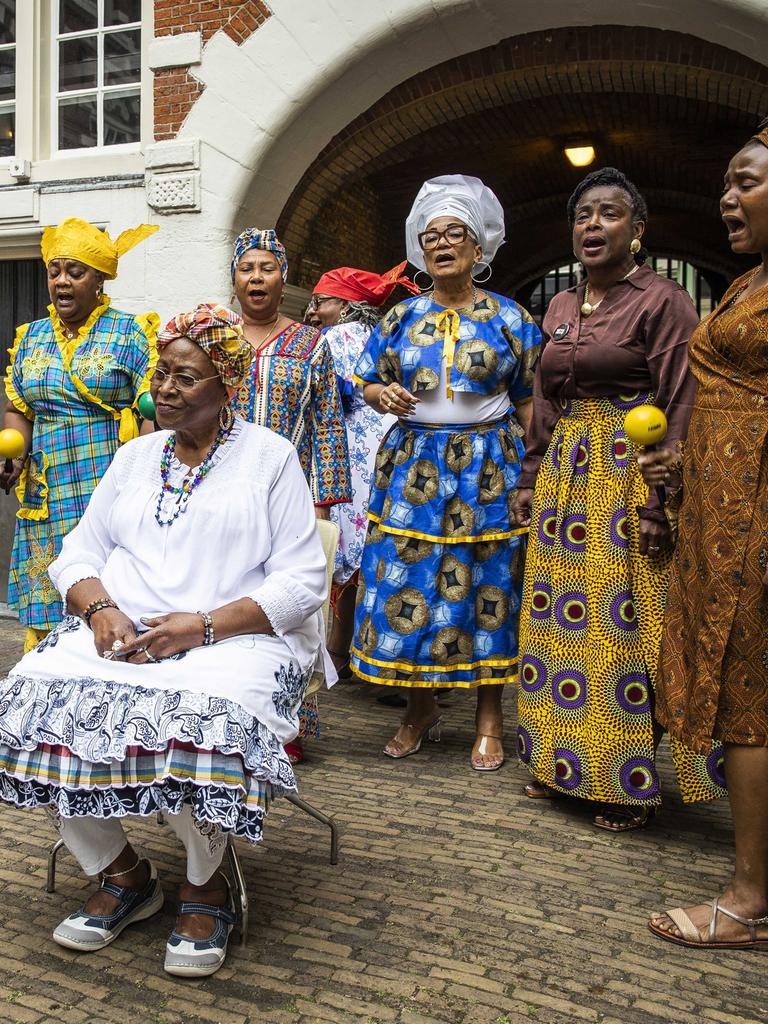 Menschen in farbenfrohen Kleidern singen in einem Hof vor einem Backsteingebäude.