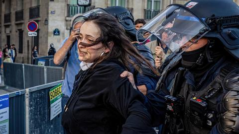Eine Frau wird in einer turbulenten Situation von einem schwer gepanzerten Polizisten abgeführt.