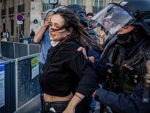 Eine Frau wird in einer turbulenten Situation von einem schwer gepanzerten Polizisten abgeführt.
