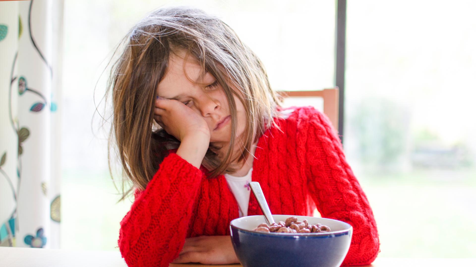 Ein müdes Kind sitzt mit geschlossenen Augen und aufgestütztem Kopf vor einer Schüssel Frühstücksflocken