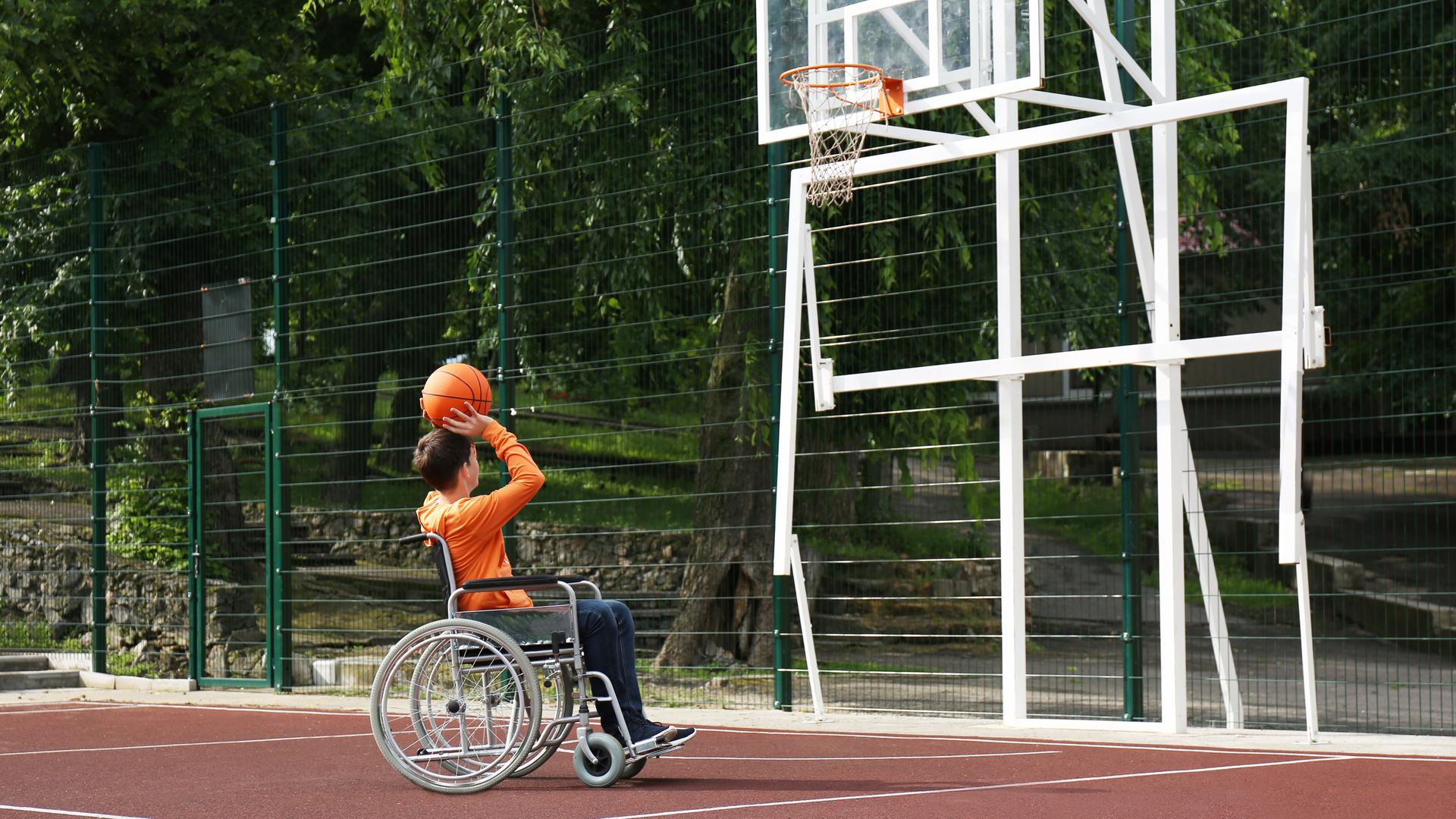 Das Bild zeigt einen Jungen im Rollstuhl, der alleine auf einem Basketballfeld einen Ball in den Korb wirft.