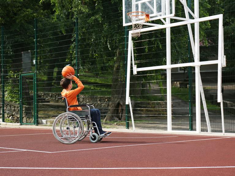 Das Bild zeigt einen Jungen im Rollstuhl, der alleine auf einem Basketballfeld einen Ball in den Korb wirft.