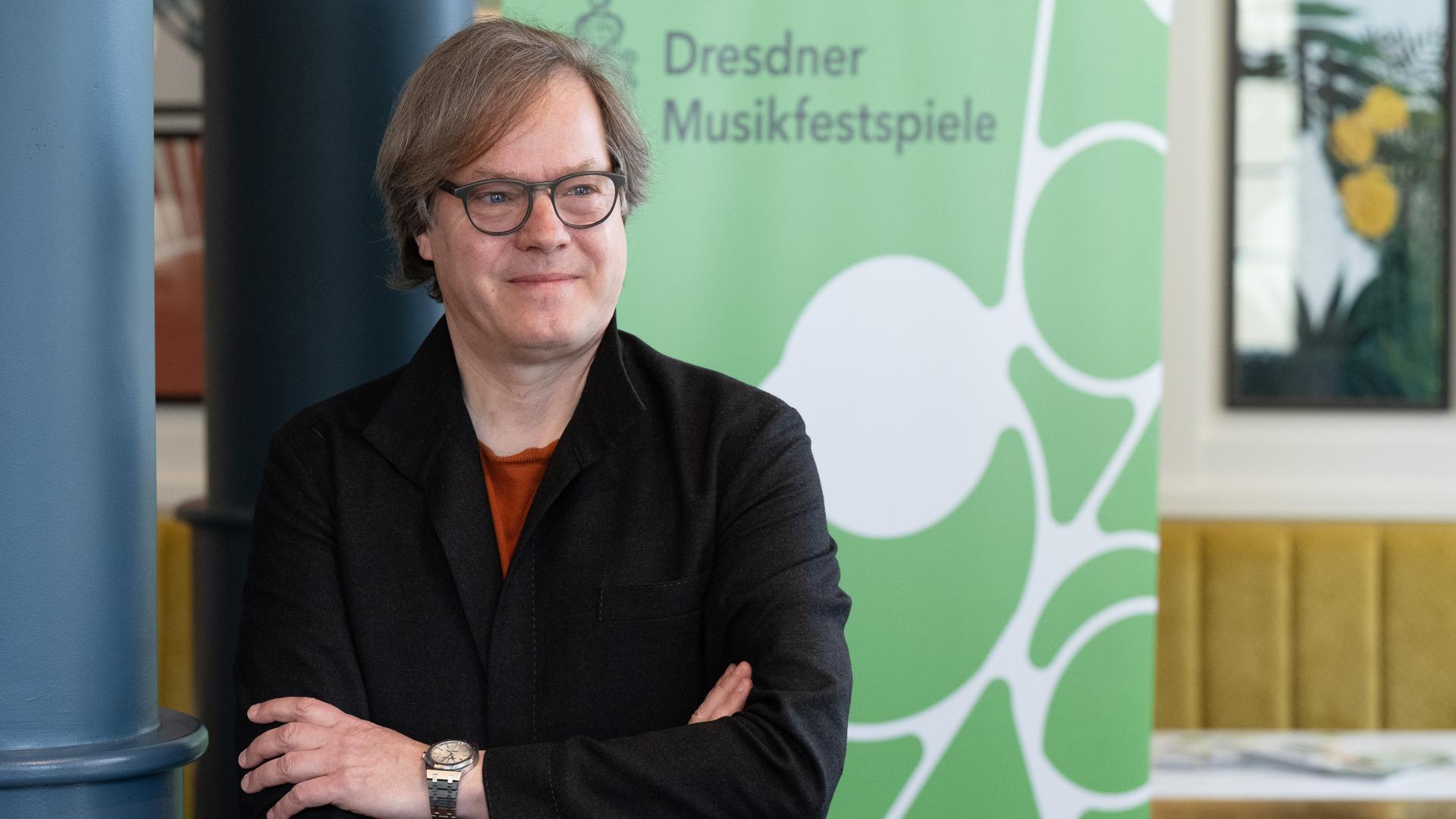 Das Bild zeigt Jan Vogler vor einem Banner mit dem Schriftzug "Dresdner Musikfestspiele". Er hält seine Arme verschränkt und hat einen zufriedenen Gesichtsausdruck. Das Bild zeigt Jan Vogler vor einem Banner mit dem Schriftzug "Dresdner Musikfestspiele". Er hält seine Arme verschränkt und hat einen zufriedenen Gesichtsausdruck.