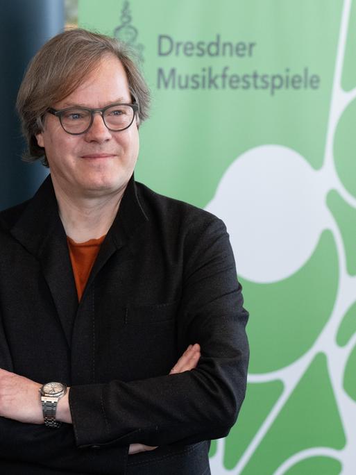 Das Bild zeigt Jan Vogler vor einem Banner mit dem Schriftzug "Dresdner Musikfestspiele". Er hält seine Arme verschränkt und hat einen zufriedenen Gesichtsausdruck.