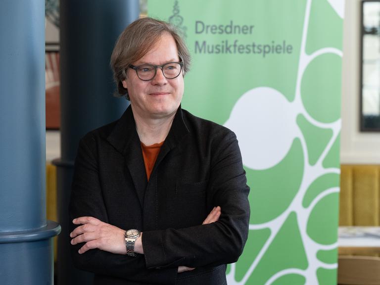 Das Bild zeigt Jan Vogler vor einem Banner mit dem Schriftzug "Dresdner Musikfestspiele". Er hält seine Arme verschränkt und hat einen zufriedenen Gesichtsausdruck.