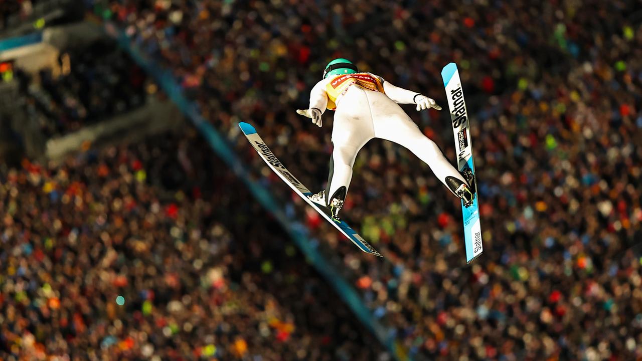 Das Bild zeigt einen Skispringer nach dem Abflug von der Schanze von oben aus schwindelerregender Höhe. Unter ihm sind Ränge voller Publikum zu sehen.