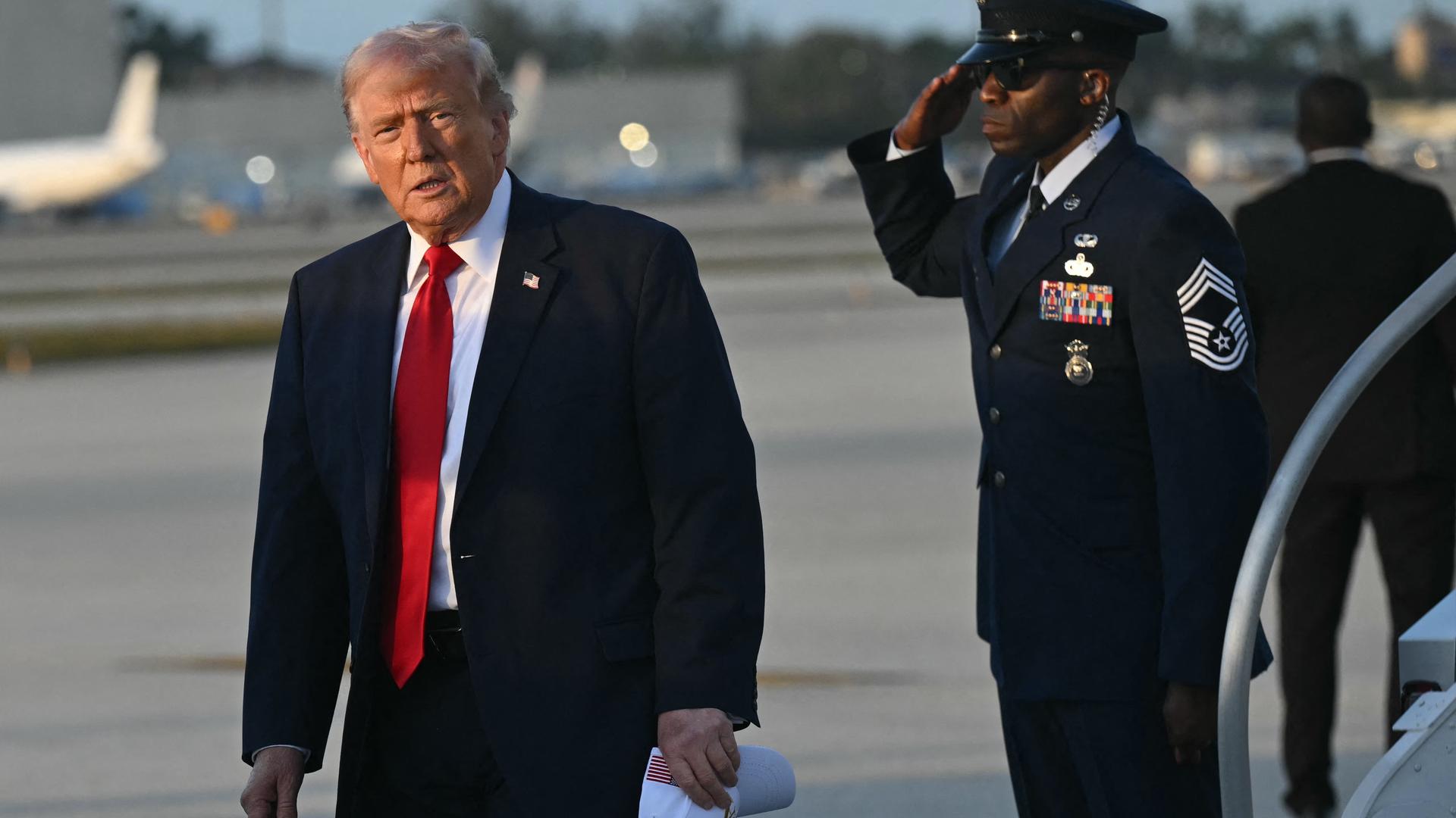 US-Präsident Trump verlässt sein Flugzeug, die Air Force One. Er blickt ernst in die Kamera, rechts von ihm salutiert ein Militärangehöriger. 