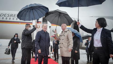 Das Foto zeigt Bundeskanzler Olaf Scholz. Er steht auf einem roten Teppich vor der Regierungsmaschine "Konrad Adenauer" auf dem Flughafen in Tokio. Mitarbeiter halten Regenschirme in den Händen.