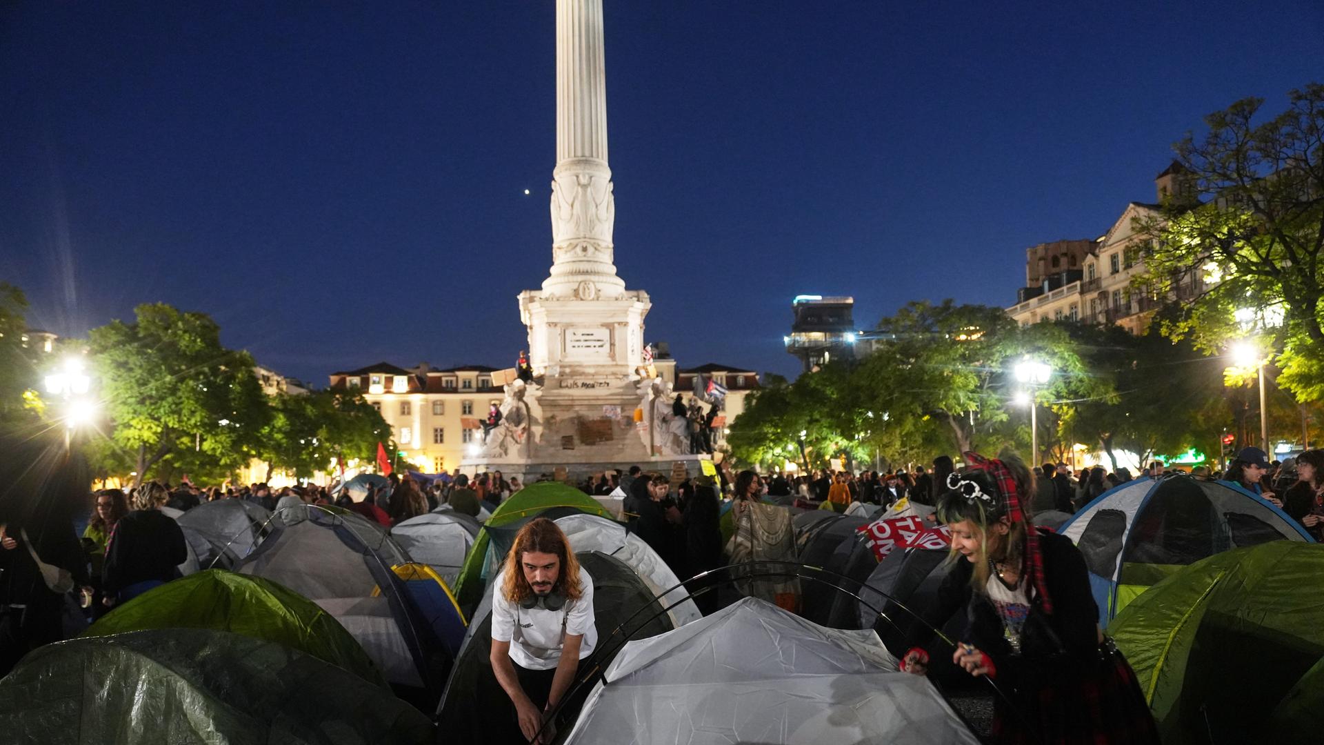 Es ist dunkel, mehrere Zelte stehen auf einem Platz in Lissabon. Zwischen den Zelten sind zahlreiche Demonstranten zu sehen. Im Hintergrund steht ein beleuchtetes Denkmal.