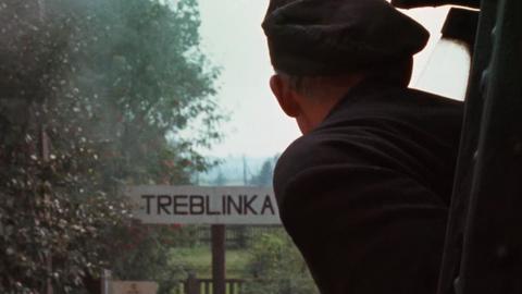 Szene aus Claude Lanzmanns Shoa. Eine Person guckt aus einem Zug, während dieser ein Schild mit der Aufschrift "Treblinka" passiert.