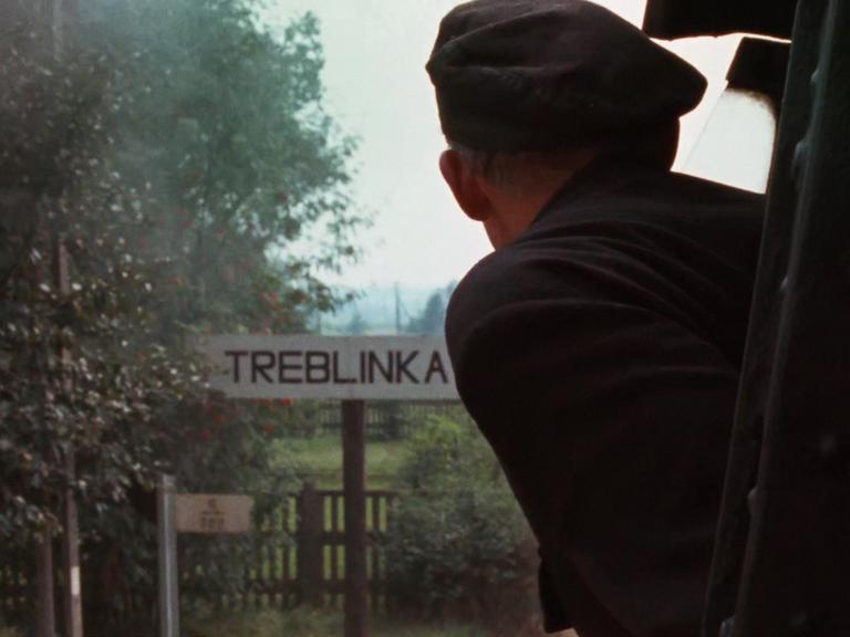 Szene aus Claude Lanzmanns Shoa. Eine Person guckt aus einem Zug, während dieser ein Schild mit der Aufschrift "Treblinka" passiert.