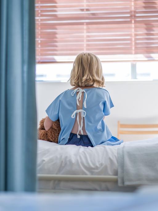 Rückansicht auf ein Kind, was auf einem Krankenhausbett sitzt, einen Teddy unter dem Arm.
