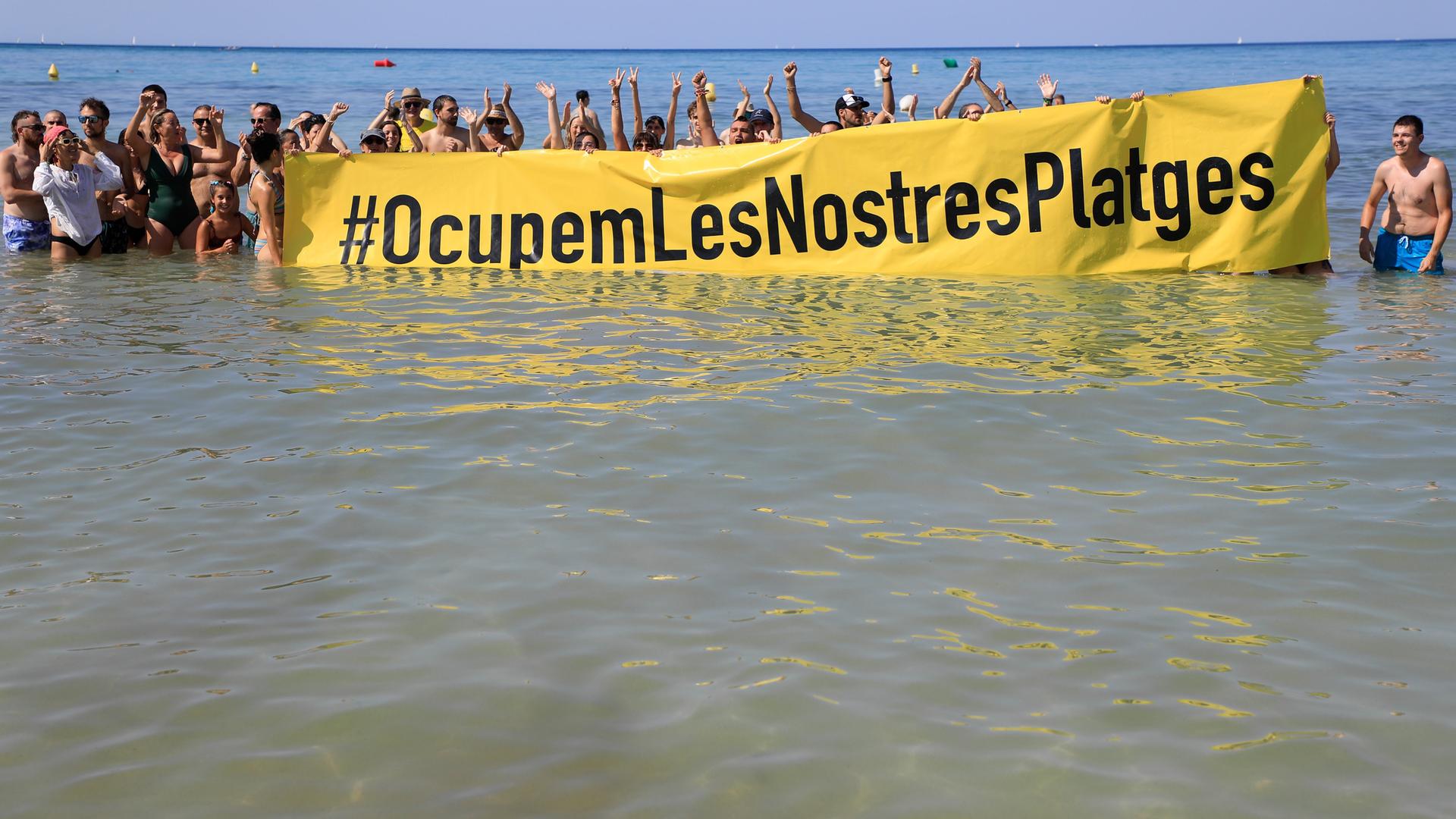 Leute halten an einem Strand auf Mallorca ein Banner "#Besetzen wir unsere Strände" aus Protest gegen den Massentourismus auf der Balearen-Insel. 