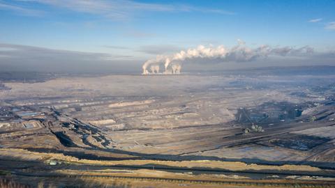 Im Vordergrund ist ein großer Braunkohletagebau zu sehen, im Hintergrund ein Kraftwerk, bei dem weiße Rauchwolken nach oben steigen.