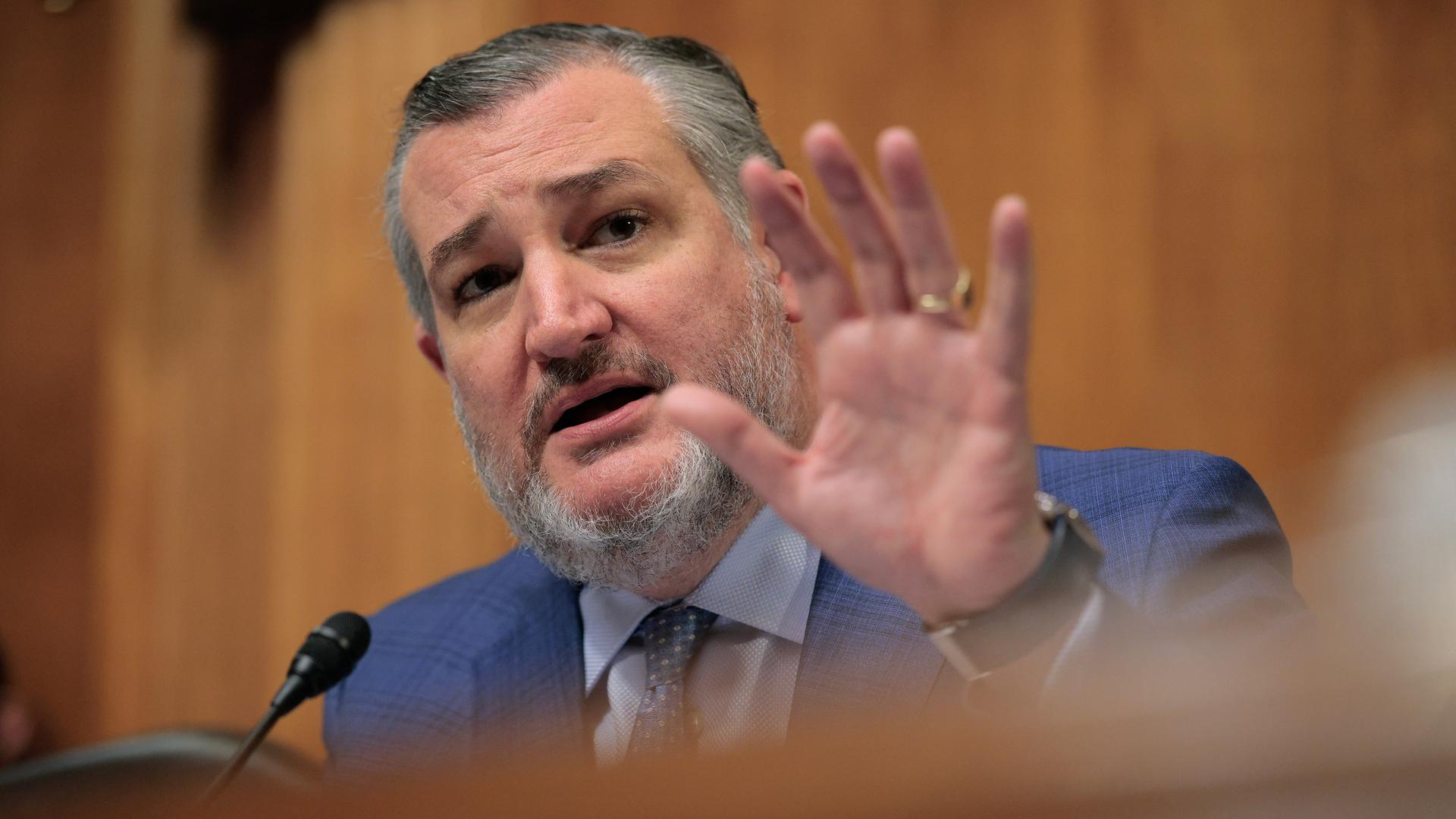 Der bärtige Cruz steht an einem Rednerpult und winkt mit der linken Hand.