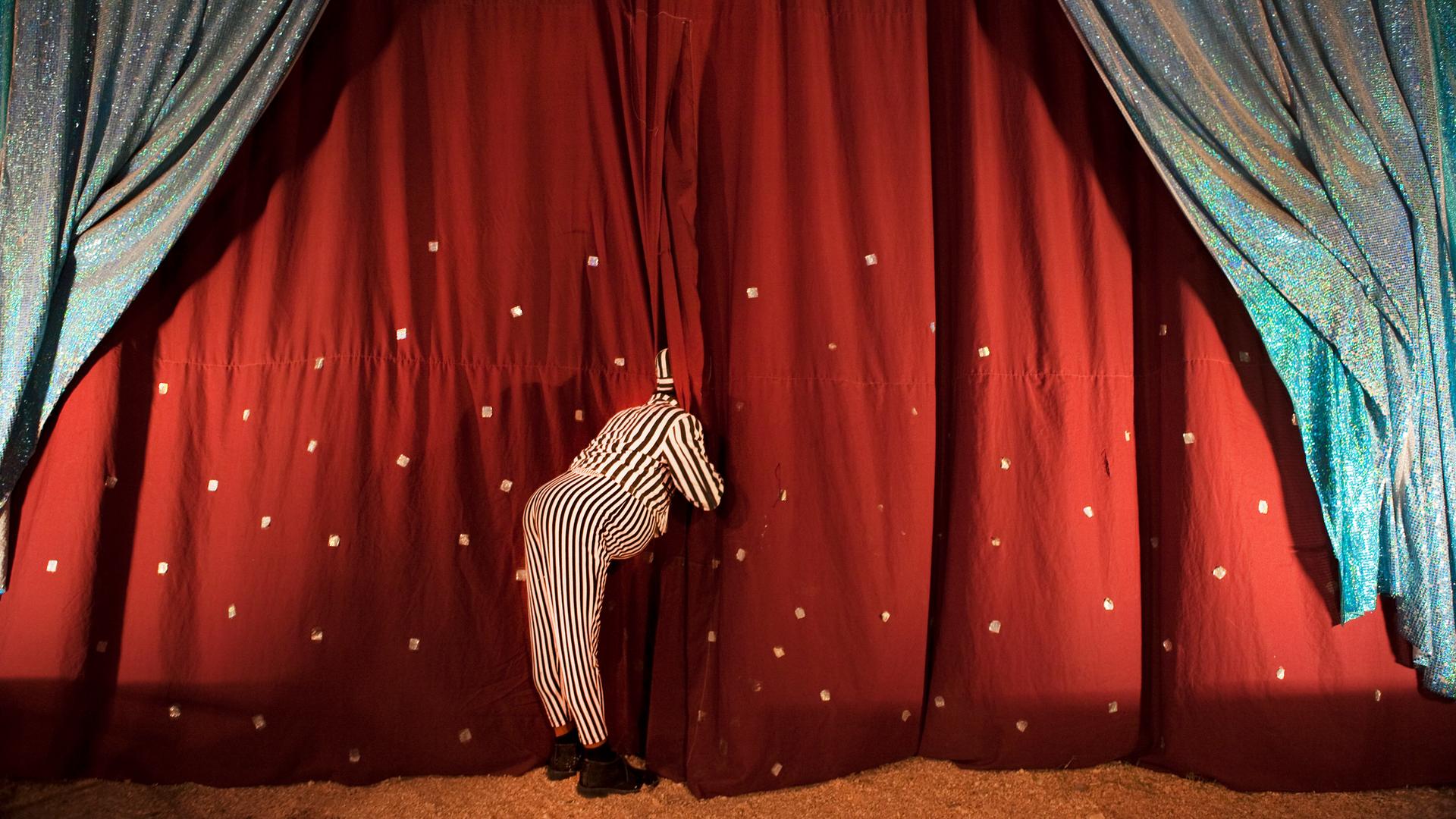 Ein Clown schaut hinter die Zirkusvorhänge, sodass sein Gesicht nicht zu sehen ist.