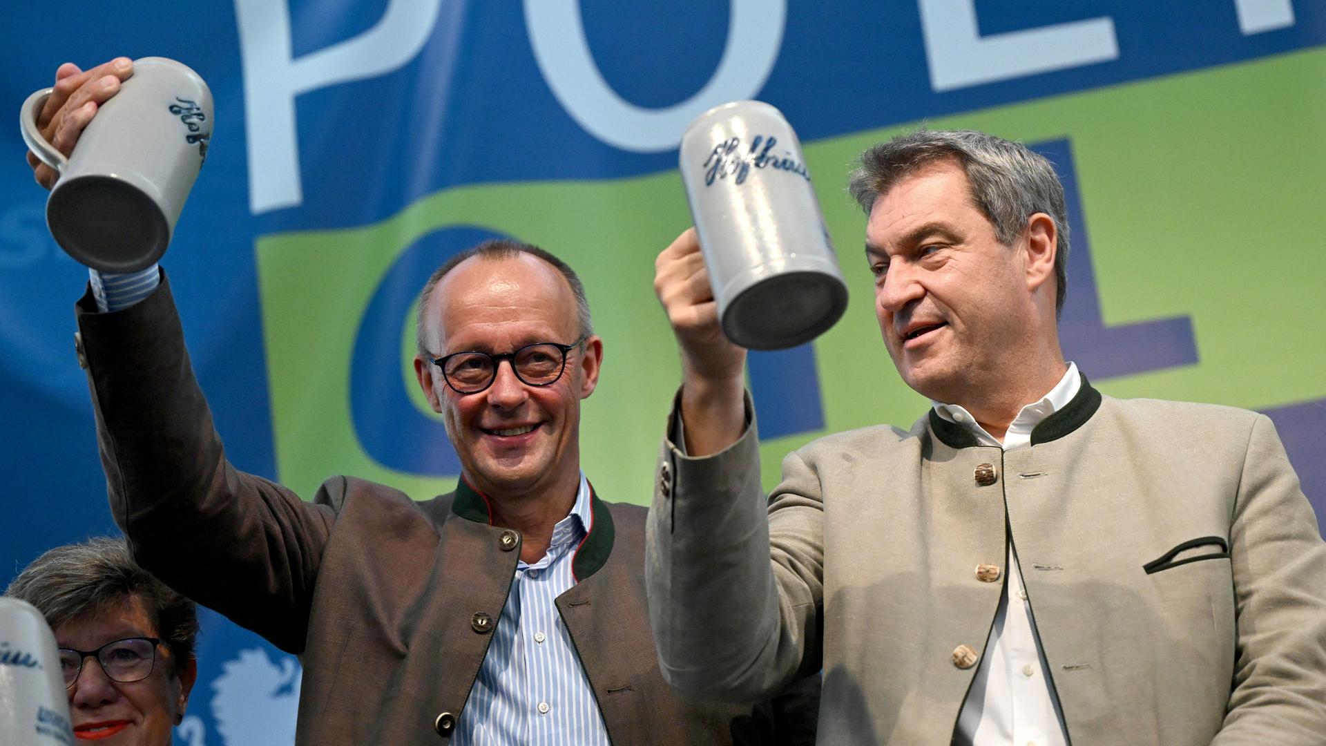 Bayern, Abensberg: Friedrich Merz (l), Bundesvorsitzender der CDU, und Markus Söder (CSU), Ministerpräsident von Bayern, prosten beim Politischen Frühschoppen Gillamoos auf der Bühne mit Bierkrügen dem Publikum zu.