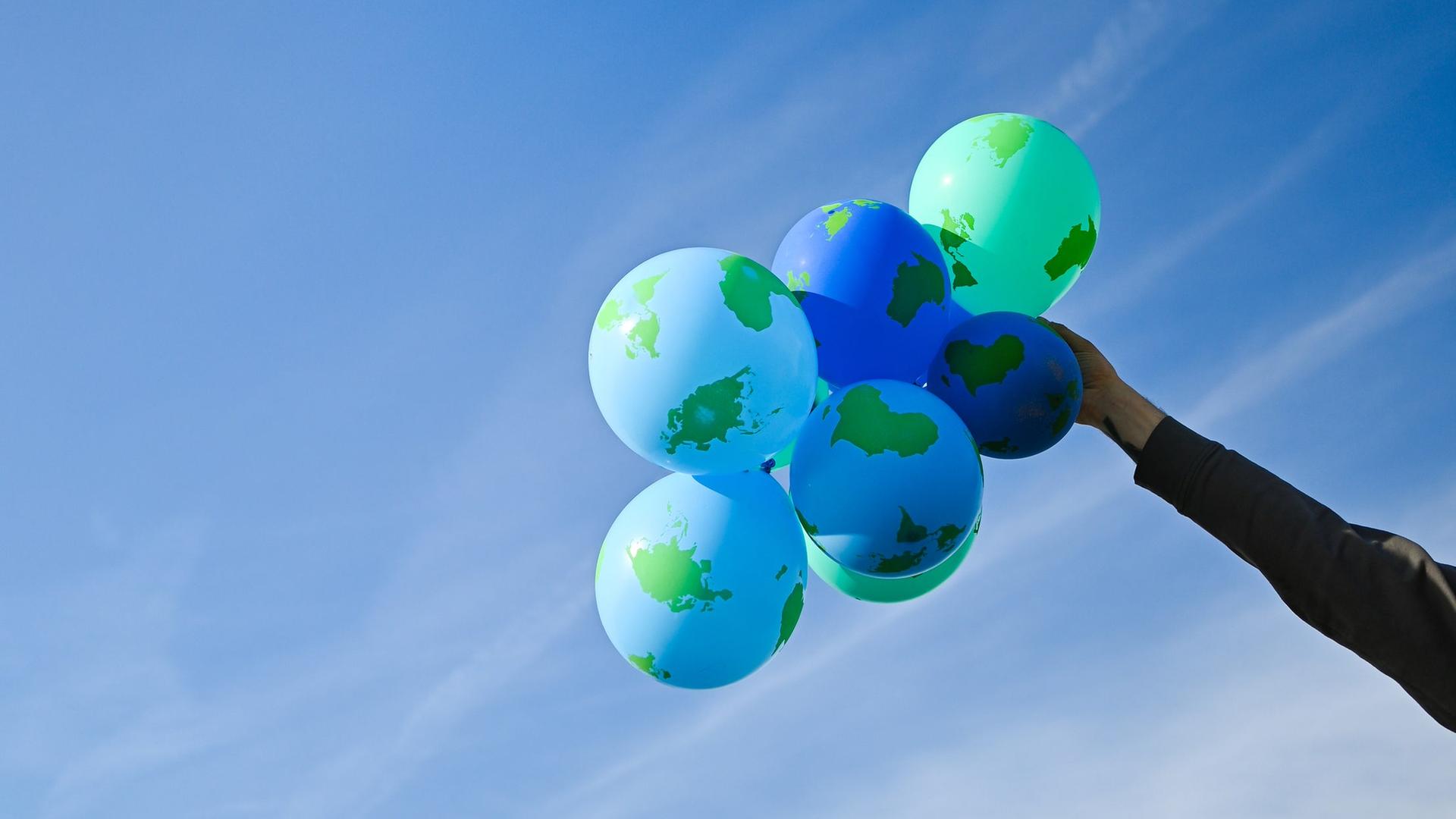Eine Hand hält einen Bündel an Weltkugel-Ballons vor blauem Himmel.