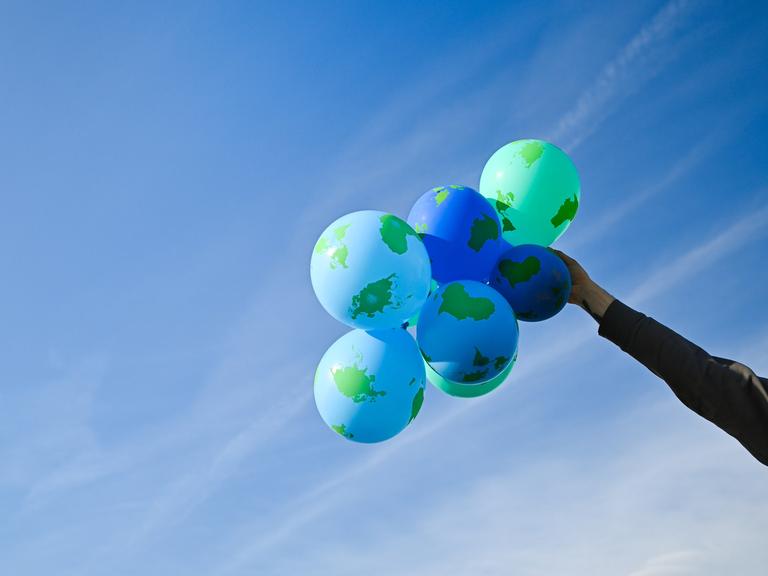 Eine Hand hält einen Bündel an Weltkugel-Ballons vor blauem Himmel.