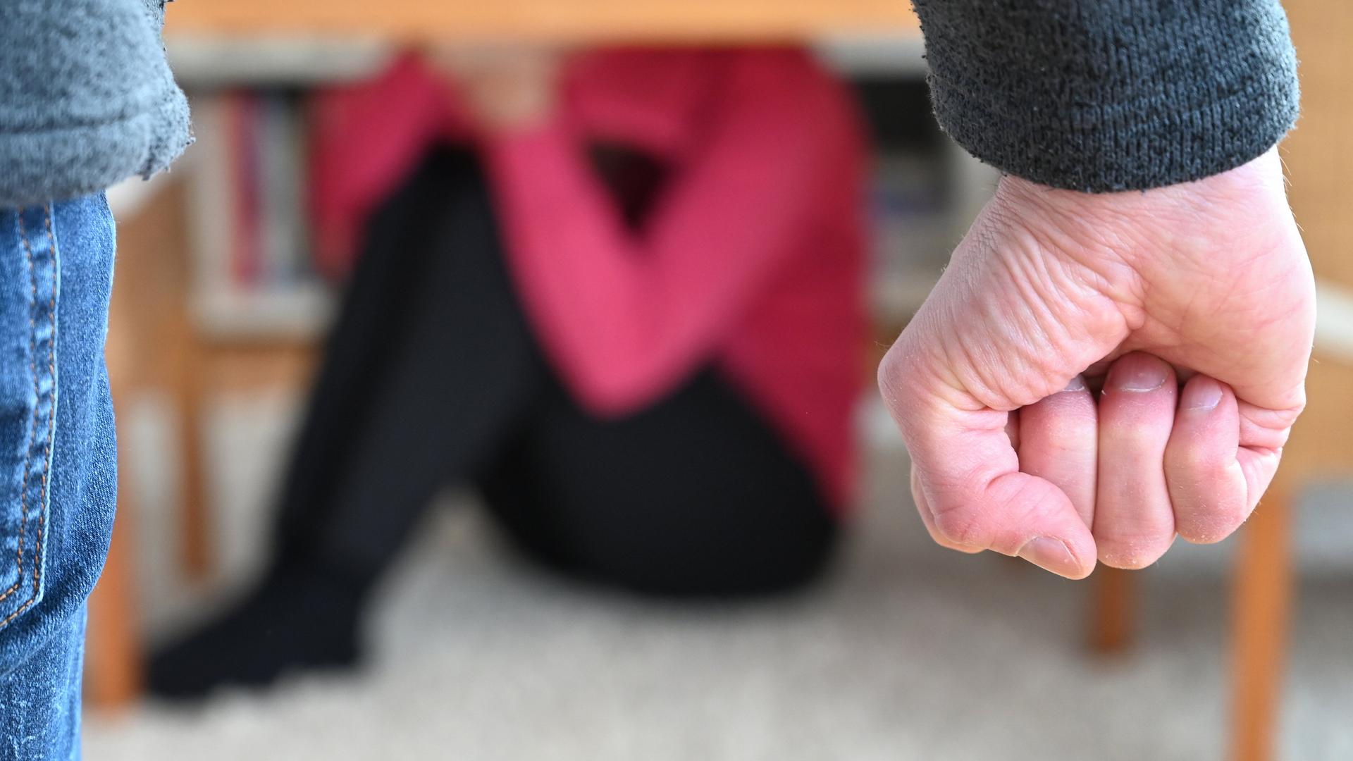 Eine Frau versucht, sich in einer gestellten Szene vor der Gewalt eines Mannes zu schützen (gestellte Szene), im Vordergrund sieht man die Faust des Mannes, im Hintergrund in der Unschärfe eine Frau unter einem Tisch.