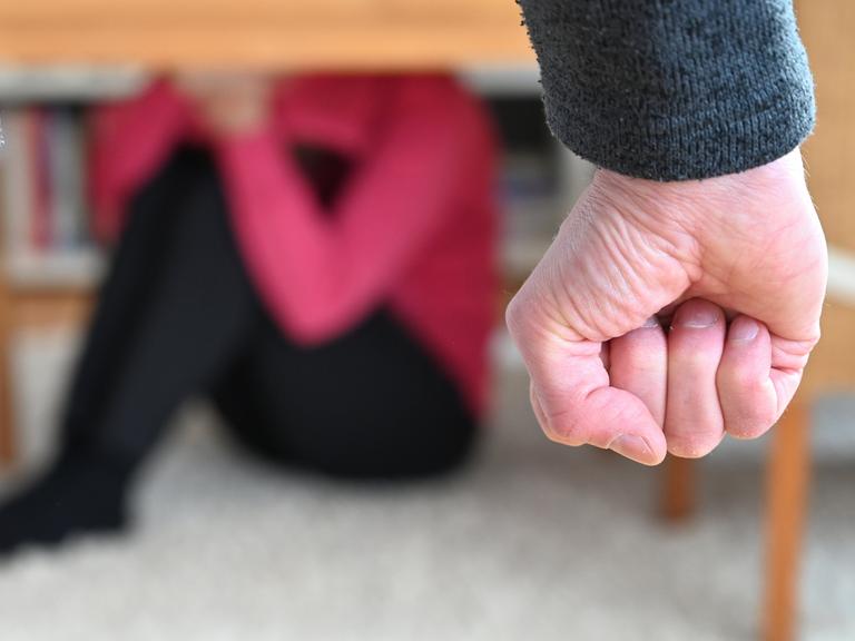 Eine Frau versucht, sich in einer gestellten Szene vor der Gewalt eines Mannes zu schützen (gestellte Szene), im Vordergrund sieht man die Faust des Mannes, im Hintergrund in der Unschärfe eine Frau unter einem Tisch.
