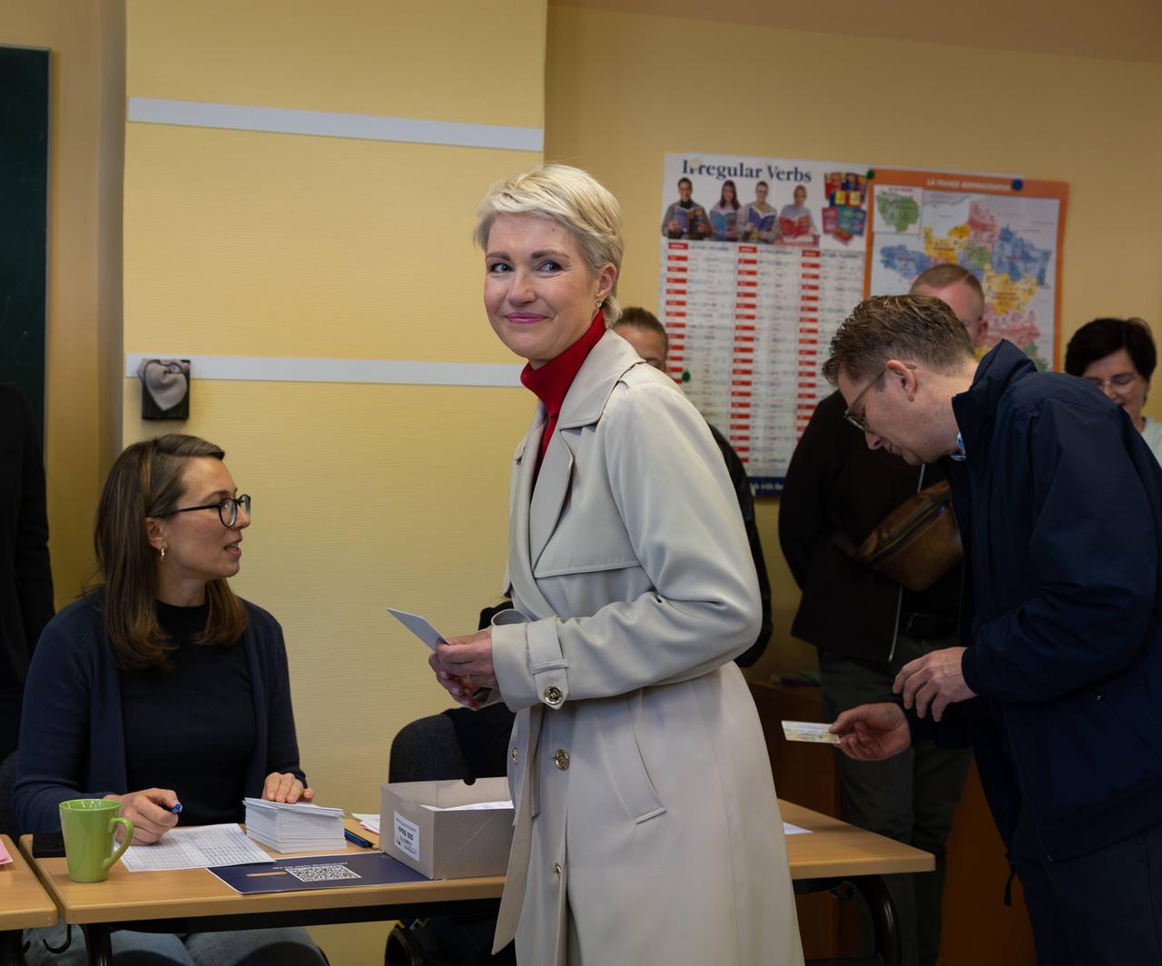 Eine Frau mit blonden kurzen Haaren steht in einem Wahllokal und hält einen Stimmzettel in der Hand.