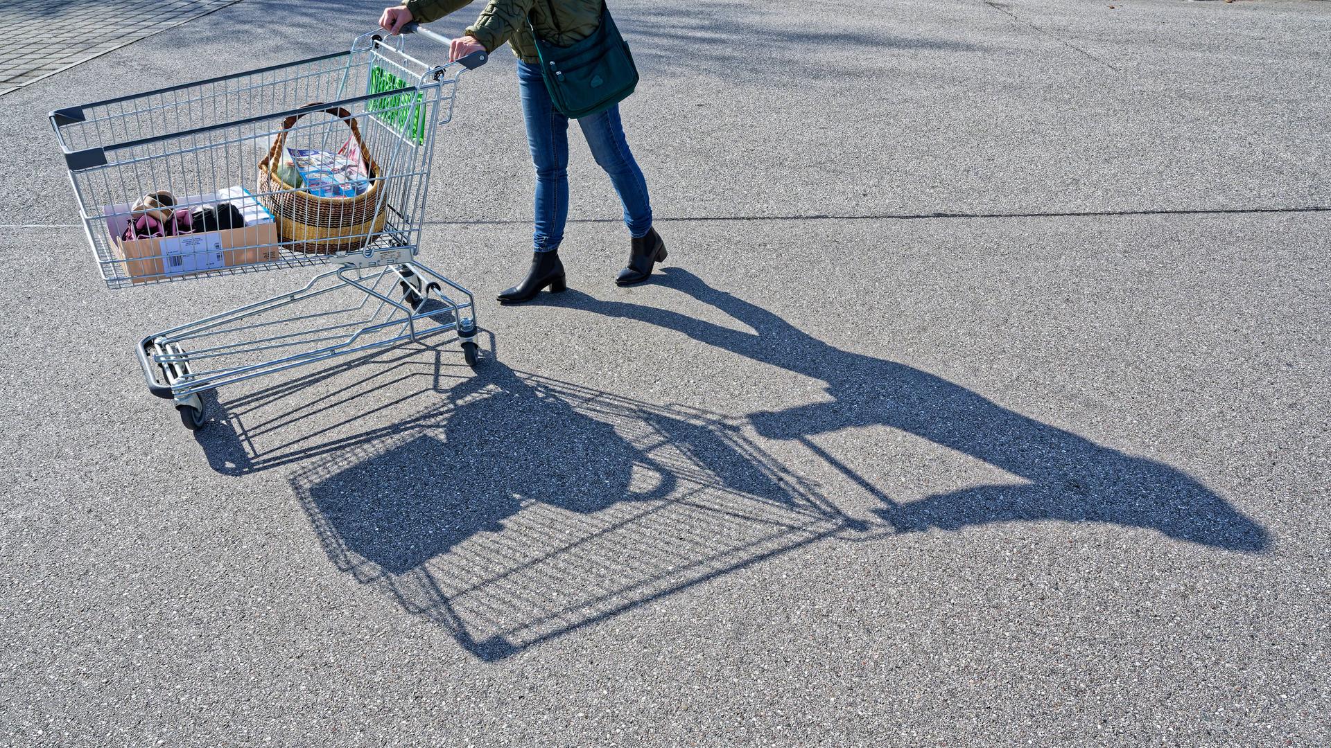 Eine Person schiebt einen halb gefüllten Einkaufswagen auf einem Supermarktparkplatz bei Sonnenschein