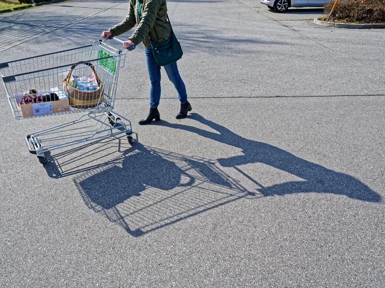 Eine Person schiebt einen halb gefüllten Einkaufswagen auf einem Supermarktparkplatz bei Sonnenschein