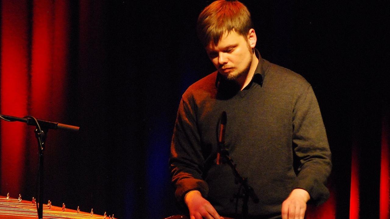 Ein junger Mann im Pullover steht auf einer rötlich-schwarz beleuchteten Bühne und spielt Zither