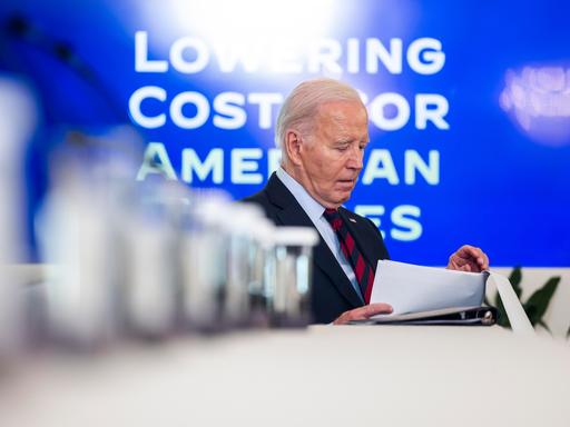 US-Präsident Joe Biden sitzt alleine an einem langen Tisch und hält ein paar Blätter Papier in der Hand.