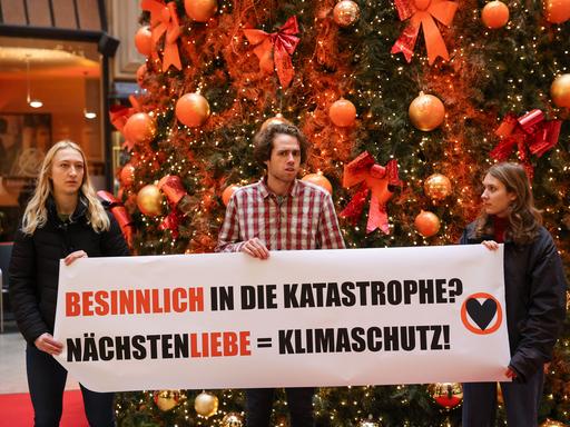 Mitglieder der Protestgruppe "Letzte Generation" stehen vor einem mit Farbe besprühten Weihnachtsbaum in der Leipziger Mädler-Passage und halten ein Plakat mit der Aufschrift: Besinnlich in die Katastrophe? Nächstenliebe = Klimaschutz.