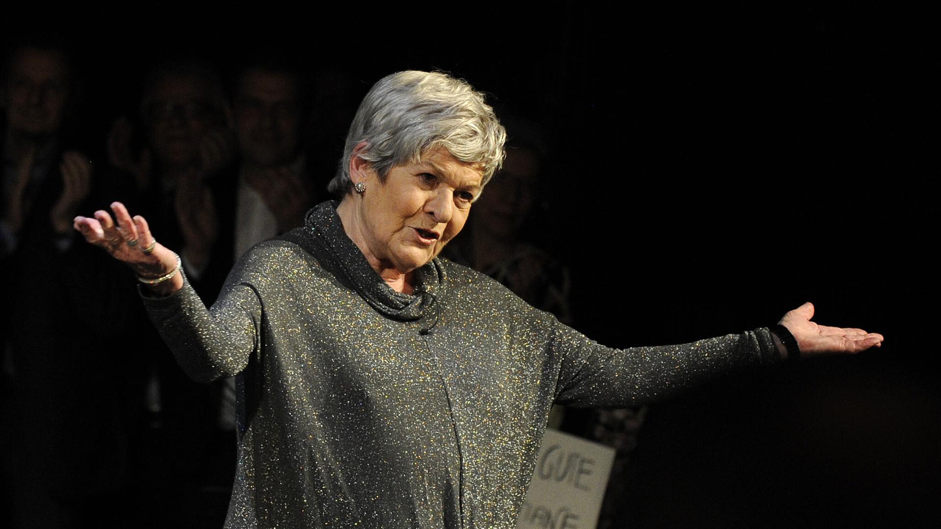 Elisabeth Orth steht auf der Bühne und breitet die Arme aus. Sie trägt ein grau-glitzerndes Oberteil. Elisabeth Orth steht auf der Bühne und breitet die Arme aus. Sie trägt ein grau-glitzerndes Oberteil.