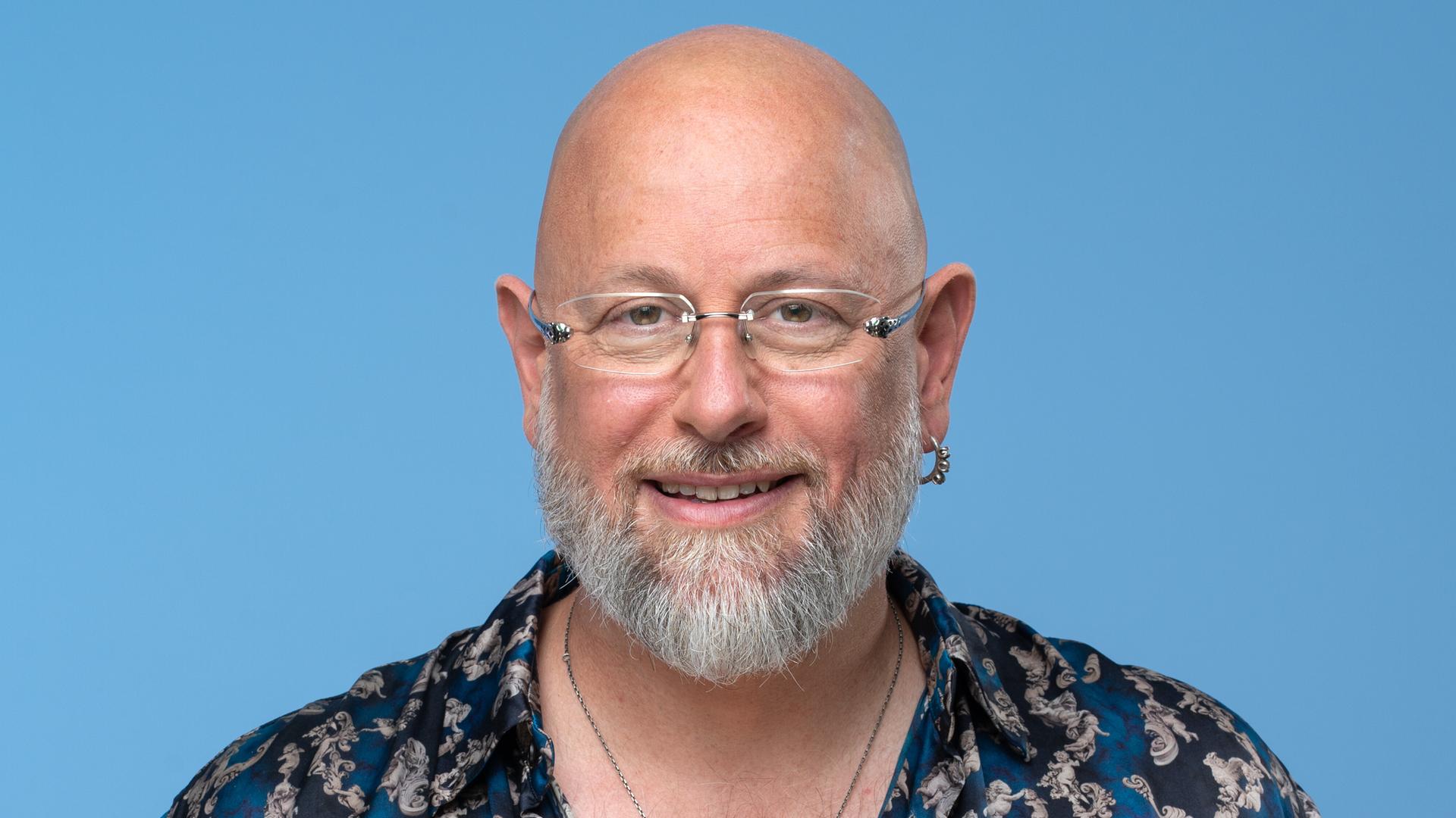 Porträt des Hypnotiseurs und Bühnenkünstlers Jan Becker. Er trägt ein blau-gemustertes Hemd und viel Schmuck und Tätowierungen. Der Hintergrund ist hell-blau. Er guckt in die Kamera. 