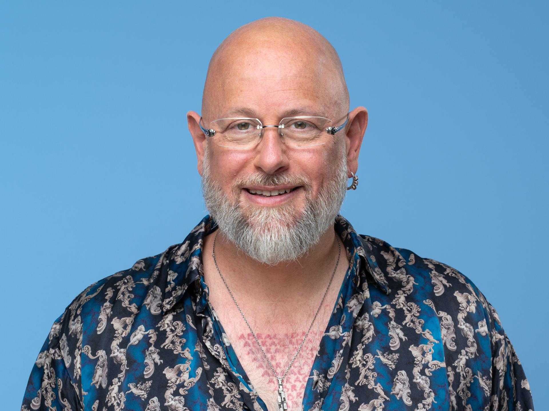 Porträt des Hypnotiseurs und Bühnenkünstlers Jan Becker. Er trägt ein blau-gemustertes Hemd und viel Schmuck und Tätowierungen. Der Hintergrund ist hell-blau. Er guckt in die Kamera. 